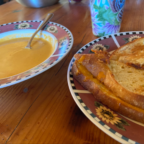 A bowl of yellow colored tomato soup and a plate of a grilled cheese sandwich.