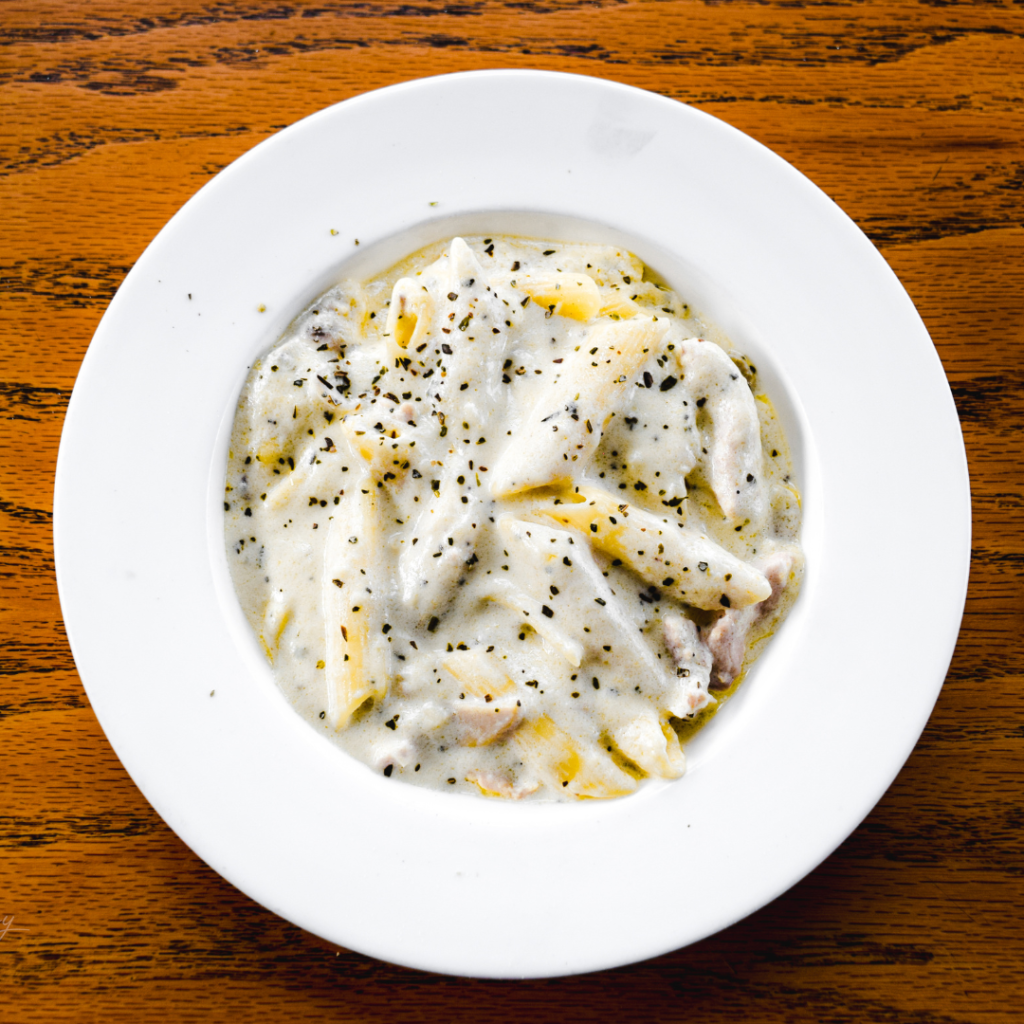 A white bowl filled with pasta, topped with a white sauce, then sprinkled with parsley.