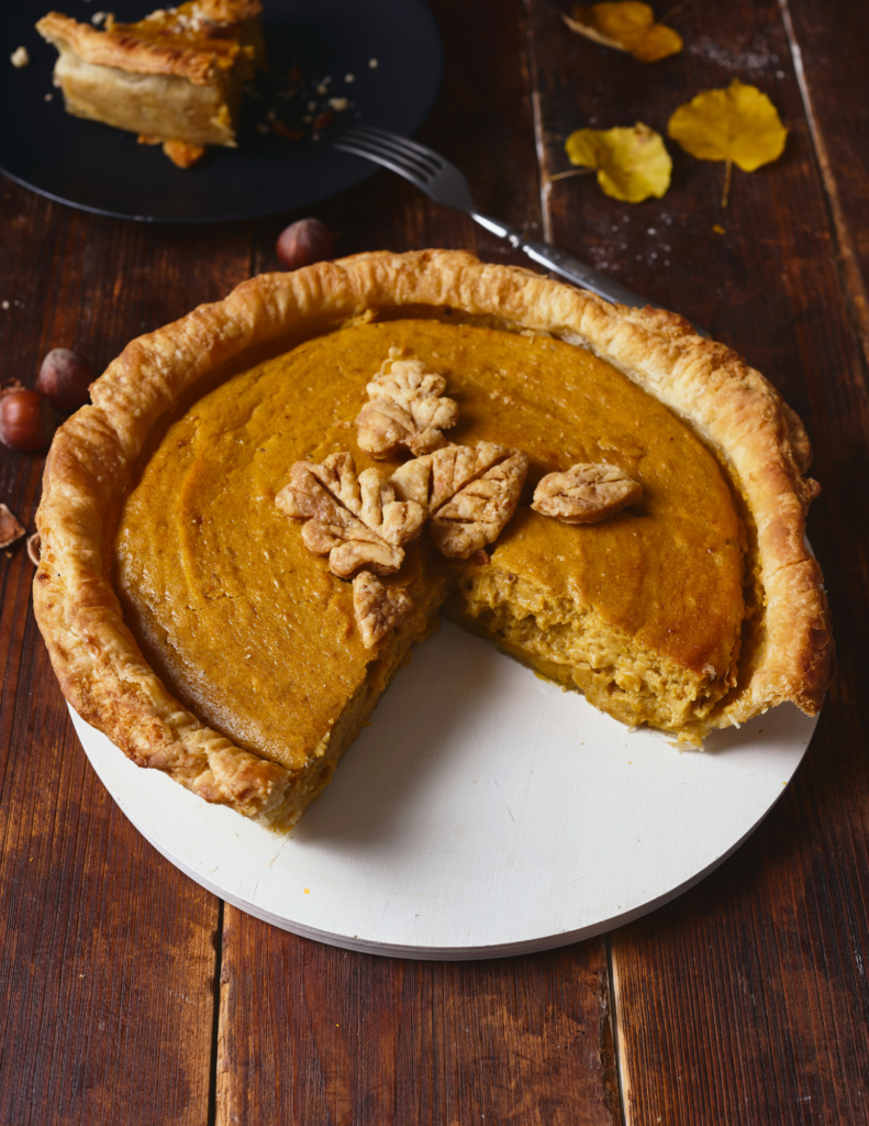 A pumpkin pie with a slice cut out on a white plate on a wooden counter.