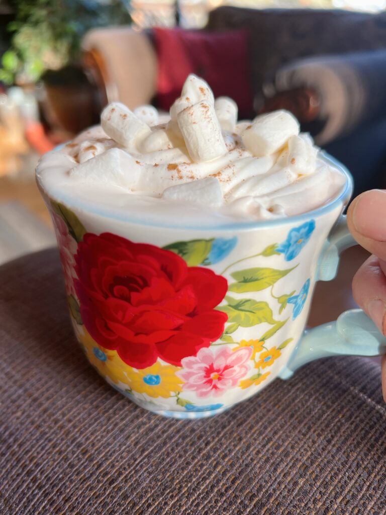 A floral coffee cup filled with hot chocolate that is topped with whipped topping and marshmallows.