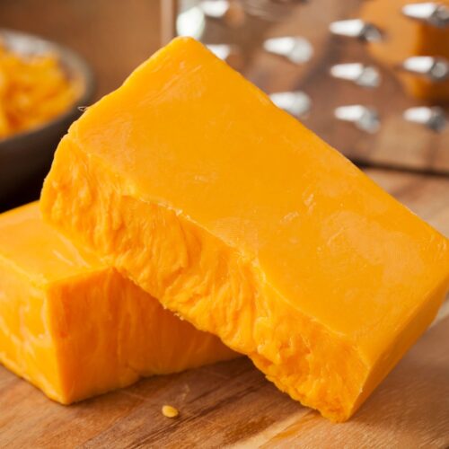 A block of cheddar cheese in front of a stainless steel cheese grater.