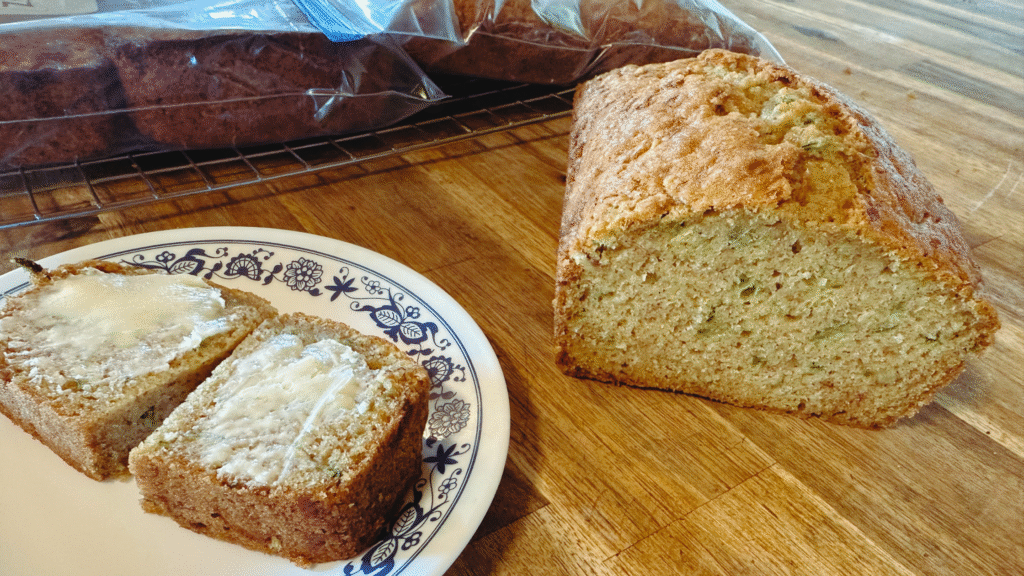 A plate with 2 pieces of zucchini bread with butter on it. The loaf is to the left and a freezer ziploc full of zucchini muffins is in the background.