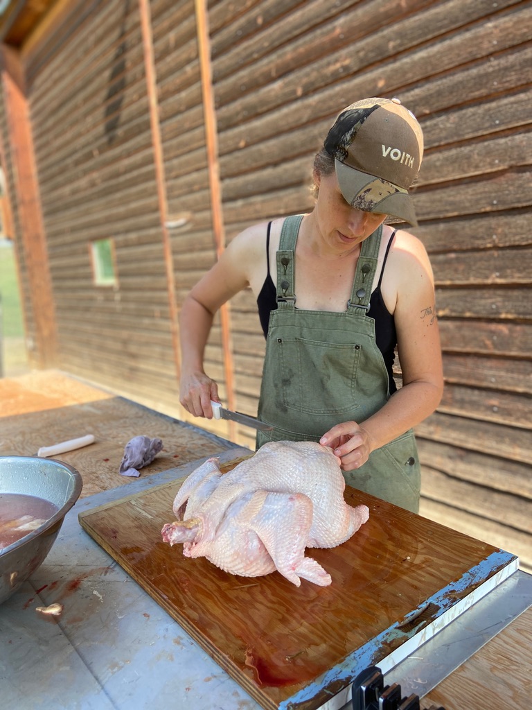 Chelsea holding a fresh whole chicken and a butcher knife about to begin parting it out.