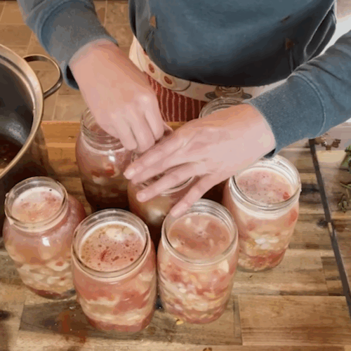 A top-down view of hands using a debubbling tool to remove air pockets from mason jars filled with vegetable soup. A large stainless steel stock pot sits to the left.
