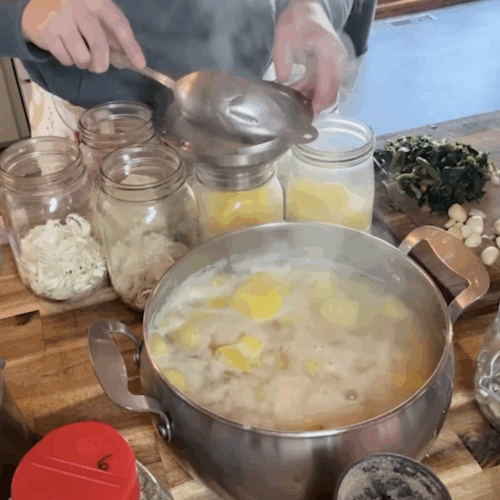 A woman using a ladle and a stainless steel canning funnel to fill mason jars with hot potatoes and broth from a large pot. Steam is rising from the food, and a food processor with chopped onions is visible on the left.