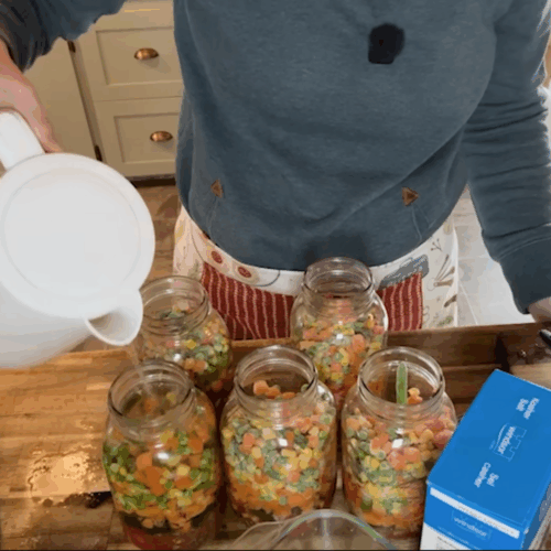 A person pouring hot water from a white electric kettle into mason jars filled with mixed vegetables like carrots, corn, and peas on a wooden kitchen counter.
