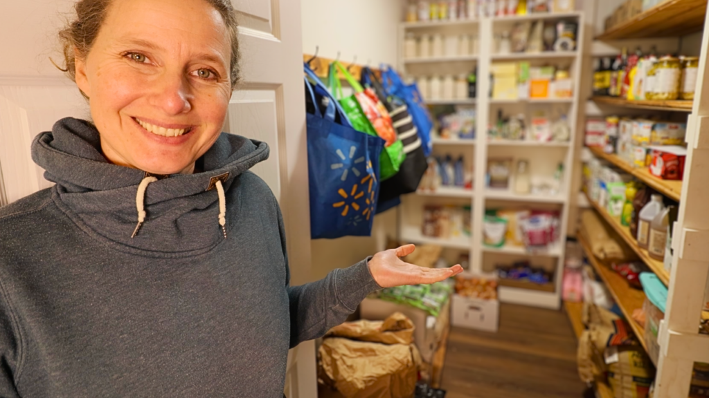 Chelsea standing in front of her well-stocked shelves, demonstrating how to easily build a 3-month pantry supply on a budget.