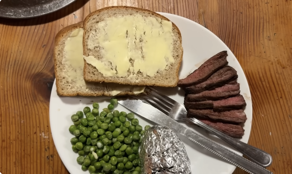 A plated dinner of sliced flank steak, peas, baked potato and buttered bread.