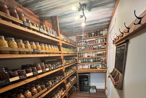 A long narrow canning pantry with shelves of canned goods on the left and an bare wall with a chalk board and hanging hooks on the right.